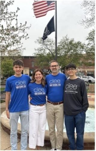 two male students and their parents standing in front of a flagpole with the American flag with blue shirts with the ciee logo