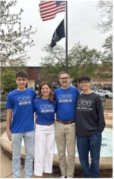 two male students and their host parents wearing with blue shirts with the ciee logo standing in front of flagpole with the American flag