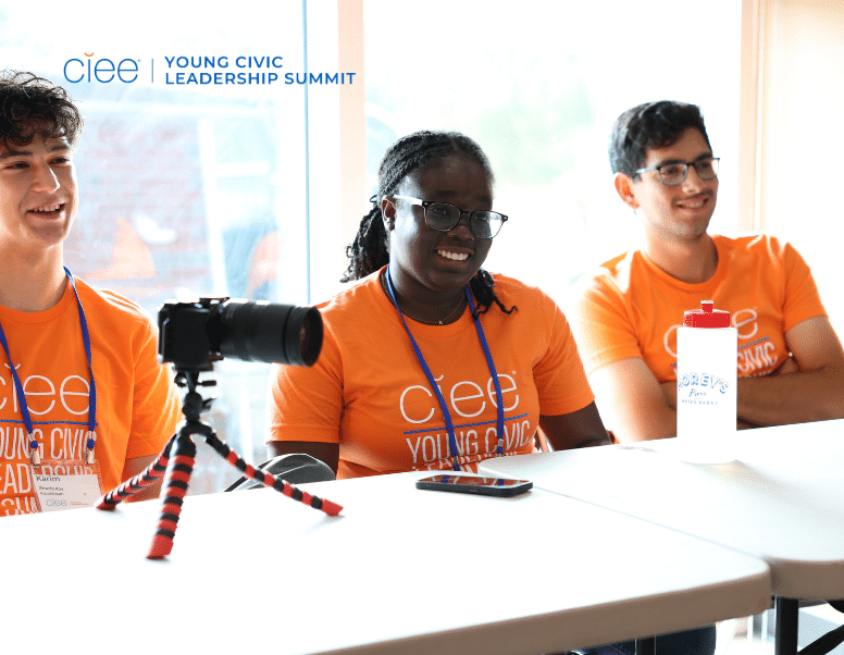 3 students sit smiling at a table wearing orange shirts that say "young civic leadership summit"